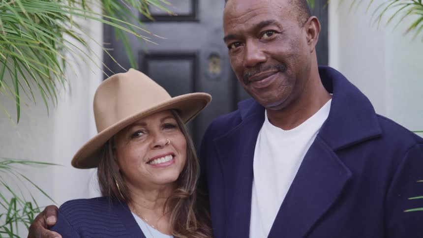 An aging man and woman stand in front of a house smiling at one another before they both turn to smile into the camera