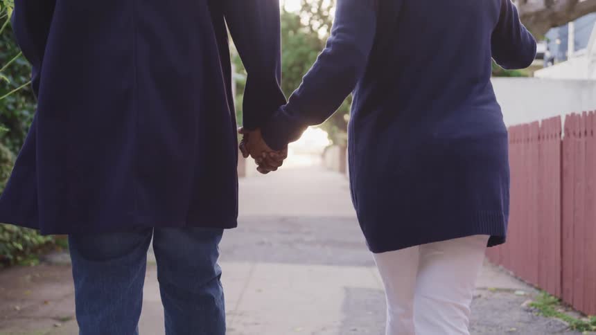 Black man with a black overcoat jeans and white woman with white pants and a blue overcoat hold hands and walk down the street