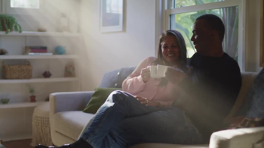 Black man with a dark blue sweater jeans and white woman with a pink shirt sitting on a sofa clinks tea cups and look into each others eyes happily