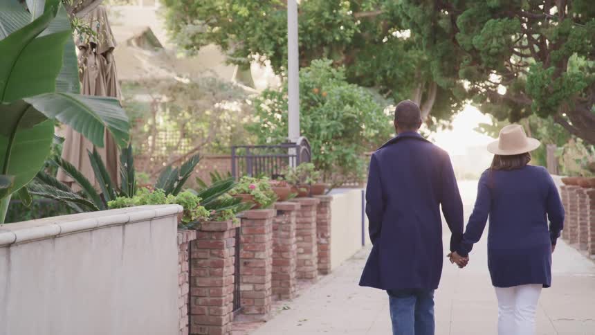 A man and woman hold hands as they walk through a neighborhood on a sunny day