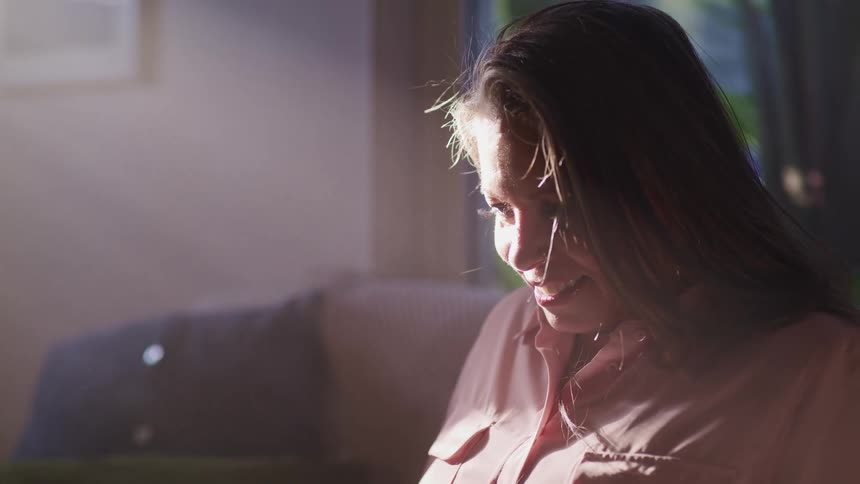 Woman with a pink shirt sits on a couch and looks down while the sun shines on her face