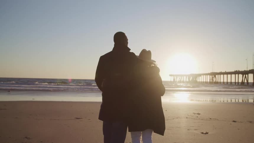 Black man with a black overcoat puts his arm around white womans shoulder and they watch sunset on the sand beach next to the sea