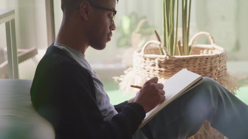 A man sits on the floor of his house and draws in a sketch book