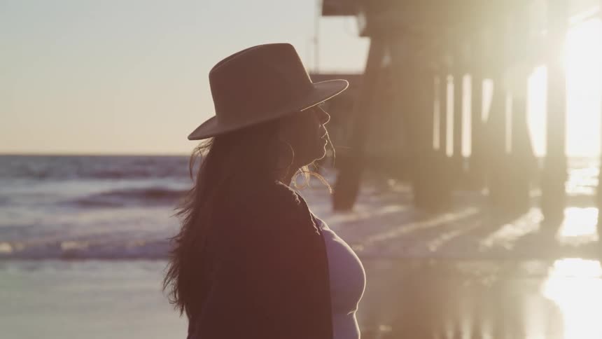 A woman in a sunhat stands lost in thought under a pier on a beach