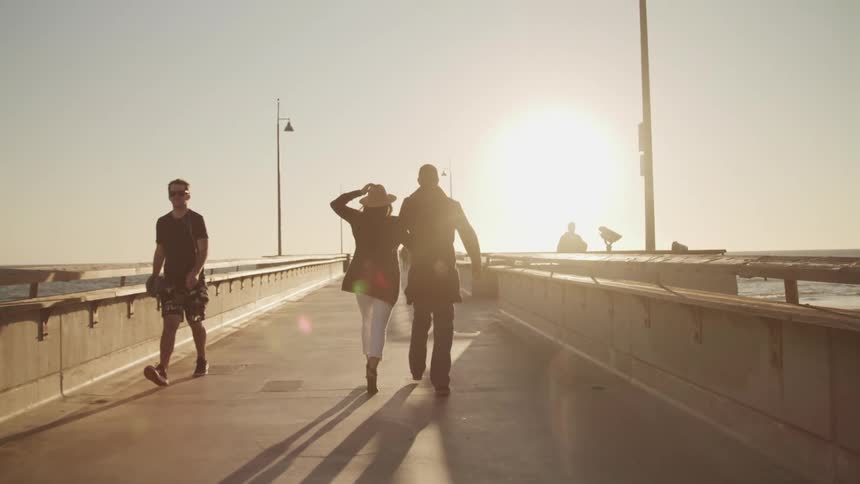 A man and a woman walking on a boardwalk with the sky and the sun in the background