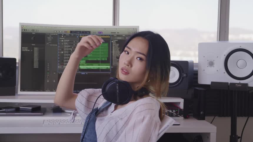 A young woman sitting at her desk surrounded by audio equipment and a computer brushes her hair off her face