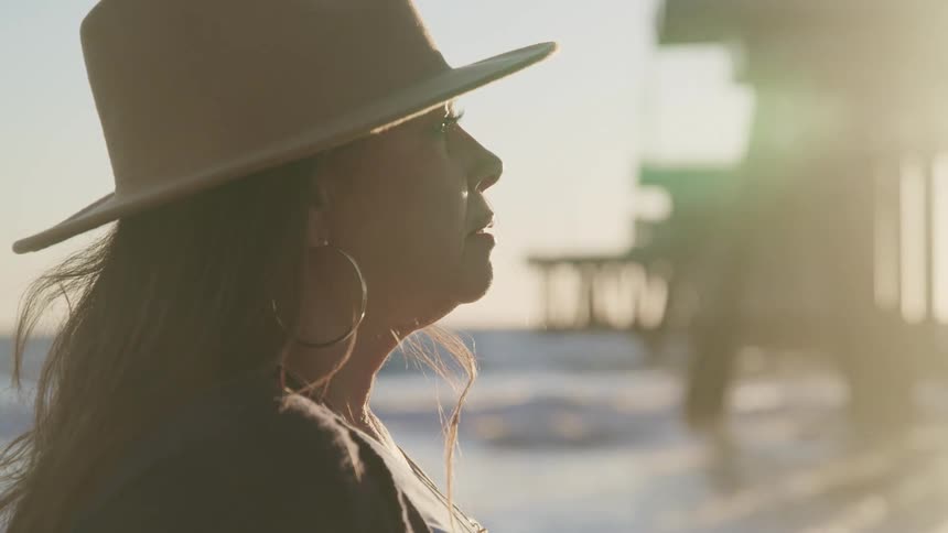 Woman wearing a brimmed hat stands on the beach near the ocean