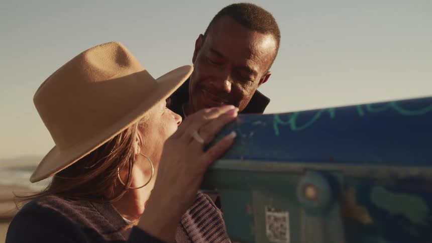 A man and woman on the beach smile at one another after a woman looks through a telescope