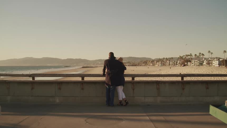 Couple reclines on the banister in a boardwalk next to the beach at sunset
