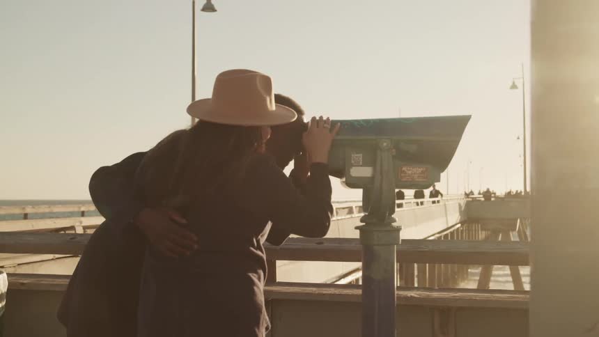 A man and woman standing on a boardwalk and looking through a telescope
