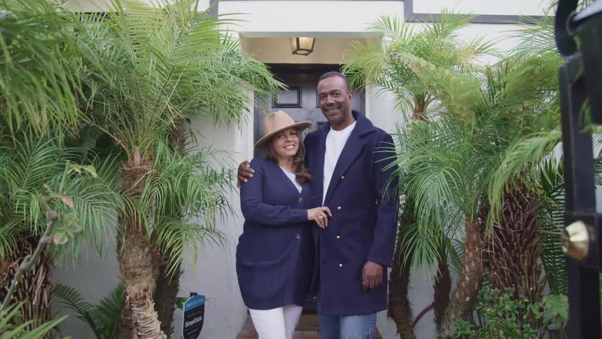 A man and woman look at each other while embracing outside of a house before turning to look into the camera