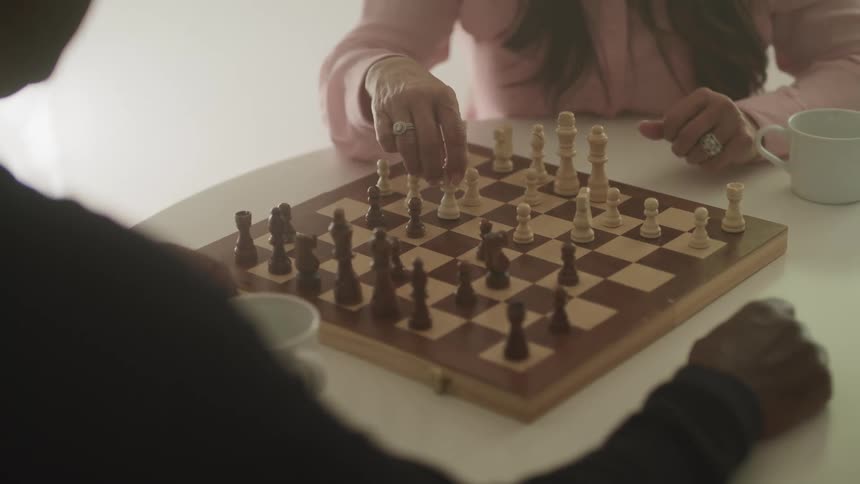 Man with a black sweater and woman with a pink sweater sits at a white table and play chess