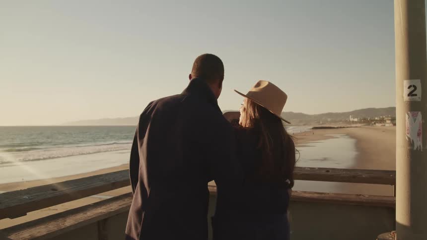 A man and woman stand and embrace on a balcony overlooking the beach