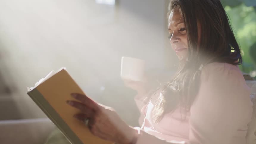 Middle aged woman with brown hair sitting on a couch and holding a cup of coffee reads a book in sunlight at home