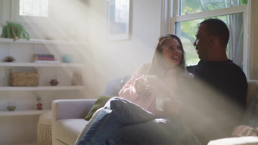 A man and woman sit together on a couch each holding a small coffee mug