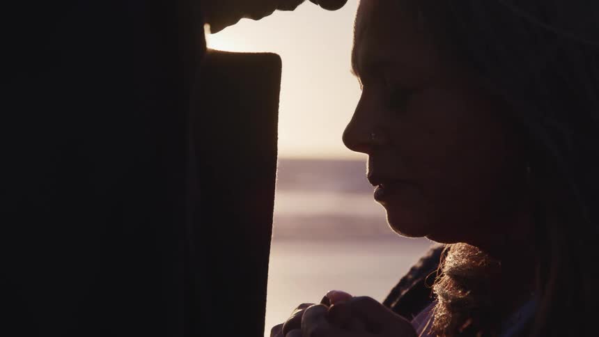 A woman standing on the shore holds a mans hands as she listens to him