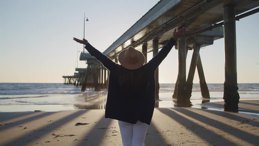 Woman stands on beach near pier supports stretching her arms up toward the clear sky