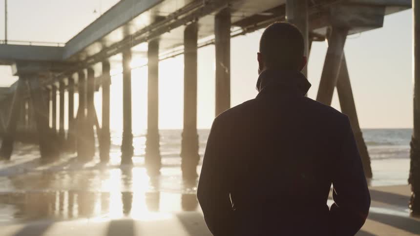 A man stands on the edge of the beach looking toward the low sun in the distant horizon