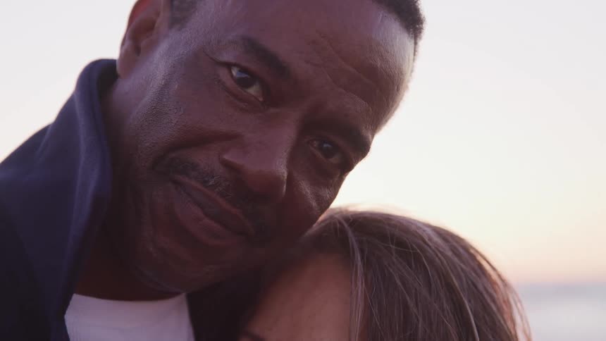 Close up of a mans face looking at the camera while holding a woman in the beach at sunset