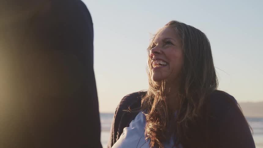 Middle aged woman with brown hair looks at mans eyes and laugh together on the beach next to the sea at sunset