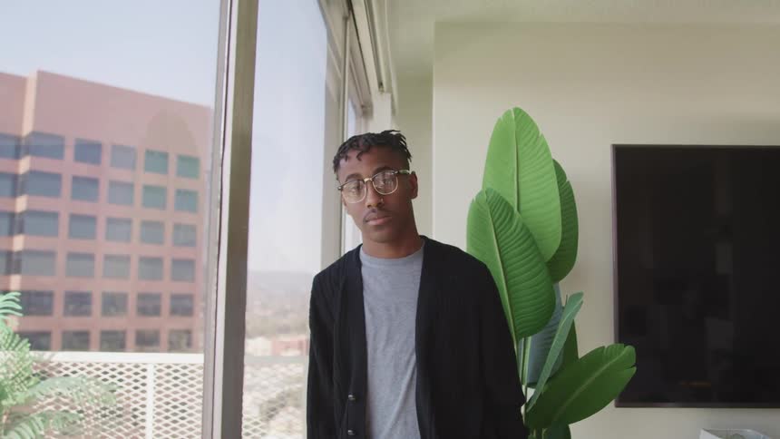 Black man with glasses leans against a window and looks at the camera in an apartment