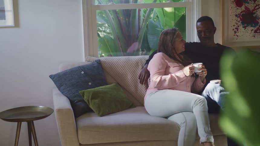 A middle aged couple sit together on a living room sofa as they drink coffee