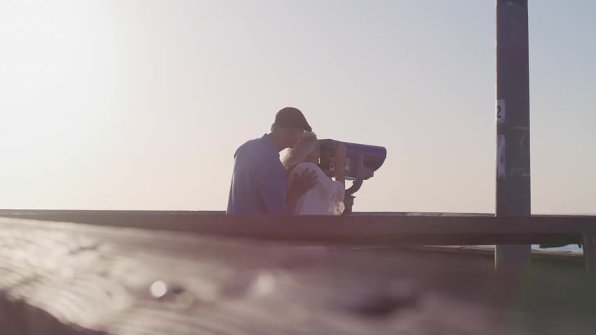 A man rests his hands on a womans shoulders as she looks through a telescope