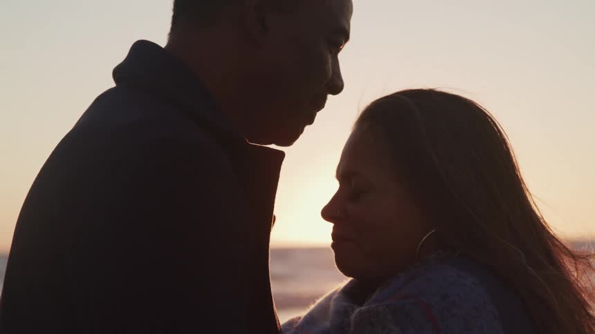 Man kisses woman on forehead as sun shining from the horizon over the ocean turns them into silhouettes