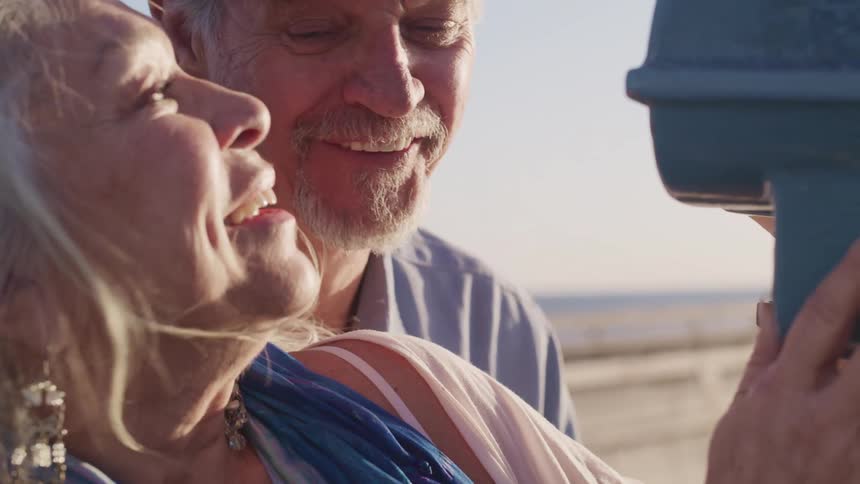 An elderly couple looks through a viewfinder together before turning to look at one another and laughing