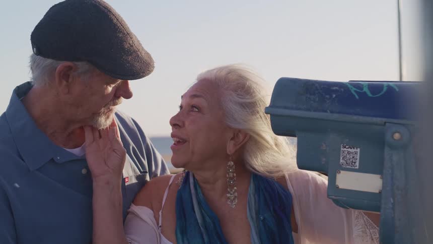 A woman rubs a mans face as she talks with him while they stand on the coast