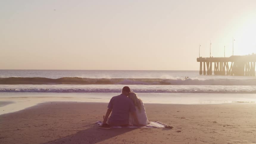 Elderly woman and man sit on the sand beach and watching sea with surfers surfing in the sea at sunset