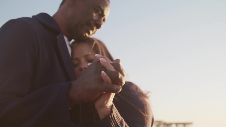 A man and woman embrace with their eyes closed while holding hands outdoors on a sunny day