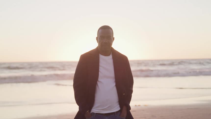 A middled aged black man stands with his back towards on the ocean on a beach at sunrise while staring into the camera