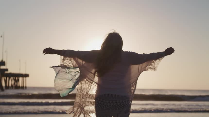 A woman wearing a sheer sweater over her clothing raises her arms joyfully while looking at the ocean waves at sunset