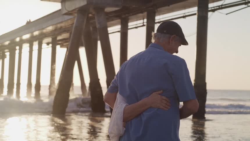 Elderly man with a cap and a blue shirt holds elderly woman wearing a shirt and walks along the beach under a pier at sunset
