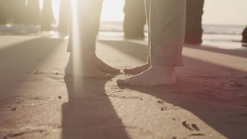 The bare feet of a pair of people standing close and facing each other on the beach during the day