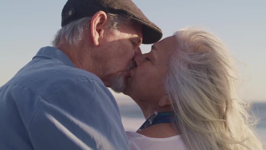 An elderly couple share a kiss while standing on a beach on a sunny day