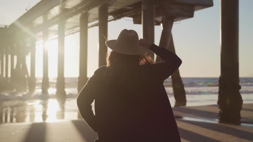 Woman stands on beach holding hat looking out toward glaring sun and ocean near pier support beams