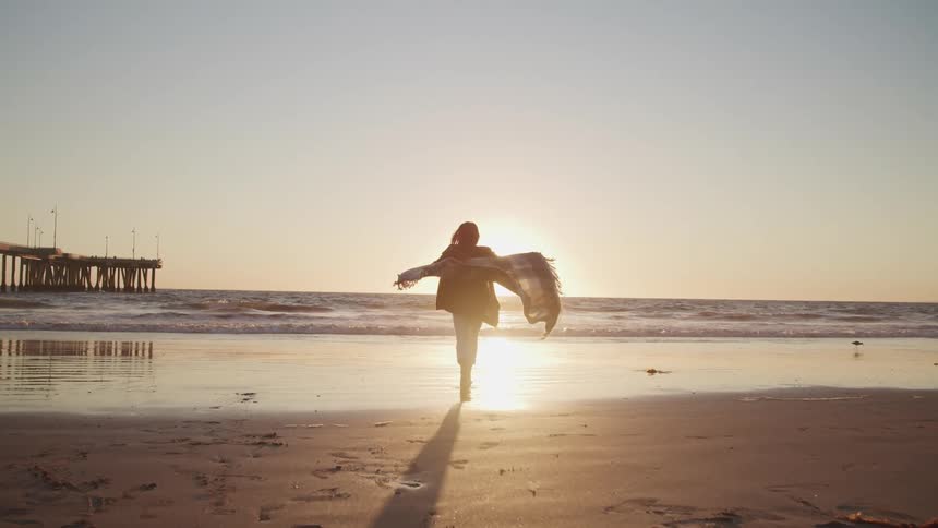 Woman runs on the sand beach with a blanket over her shoulders at sunset