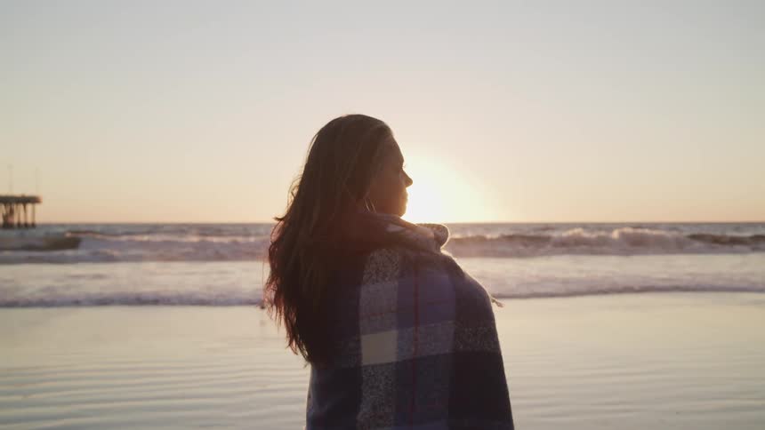 A woman standing on a beach wrapped in a blanket with the sky and the ocean in the background