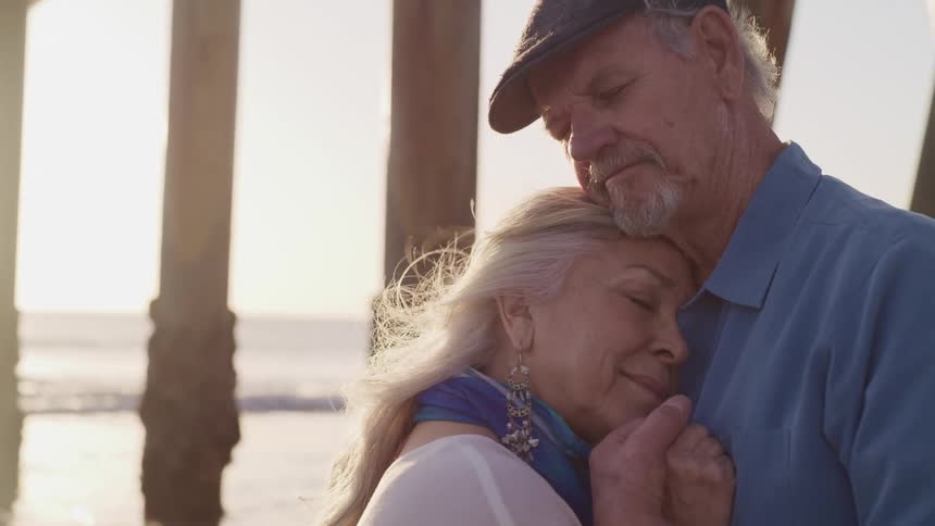 Elderly woman with a silk scarf and earrings rests her head on elderly mans chest on the beach at sunset next to the sea
