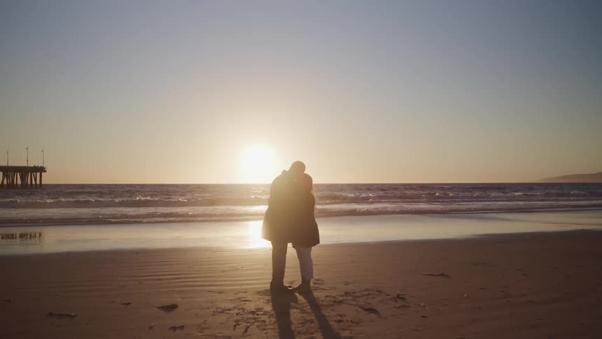 A man and woman embrace closely standing on the beach looking to the low sun of the horizon
