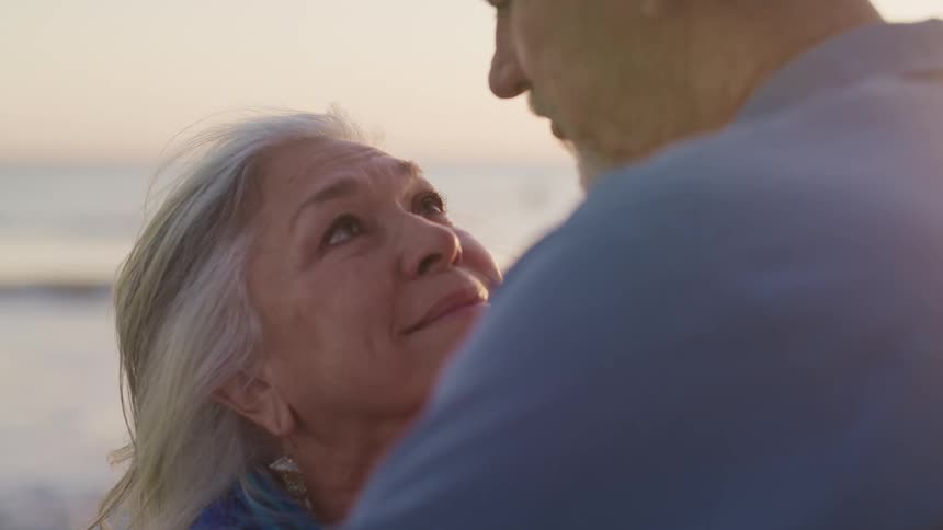 Elderly woman and man lovingly look into each others eyes on the beach at sunset