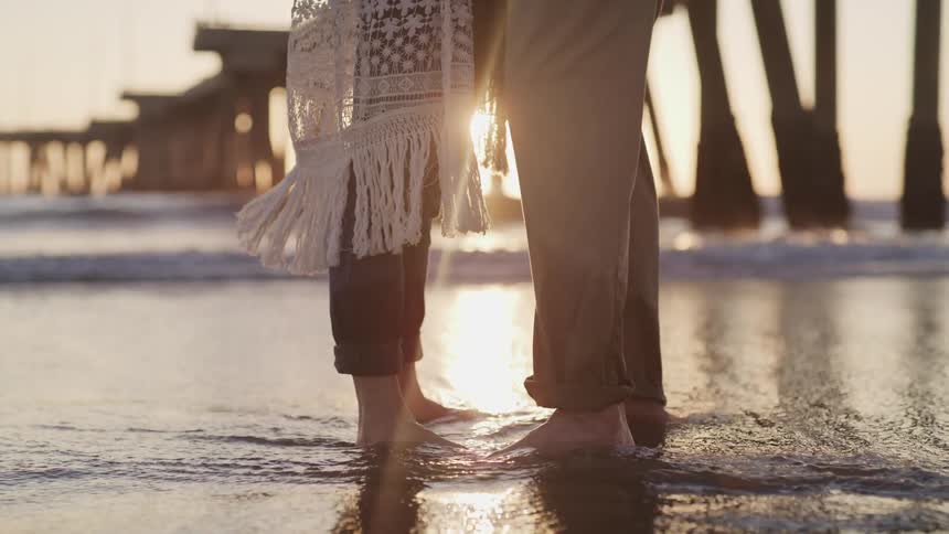 A couple stands close on the beach in front of a setting sun as the ocean water washes over their feet