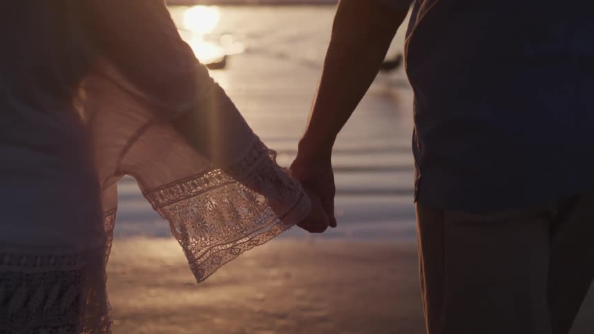 A man and woman hold hands while walking on the beach towards the ocean at sunset