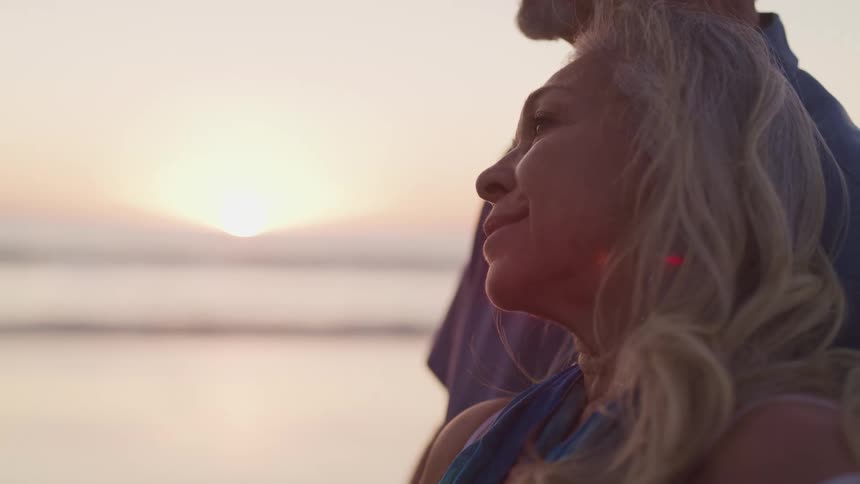 An elderly woman rests on the shoulder of an elderly man as the two stand on the beach and gaze out over the ocean as the sun peaks over the horizon