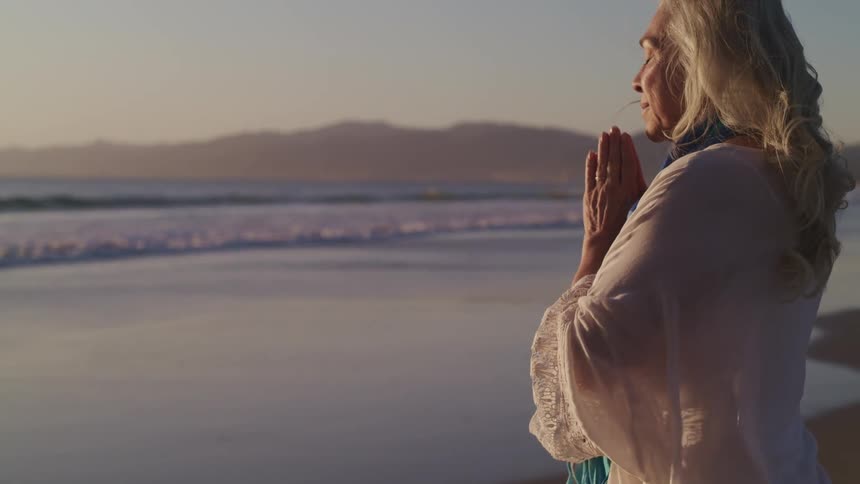 Woman puts her hands together to meditate while standing on the beach