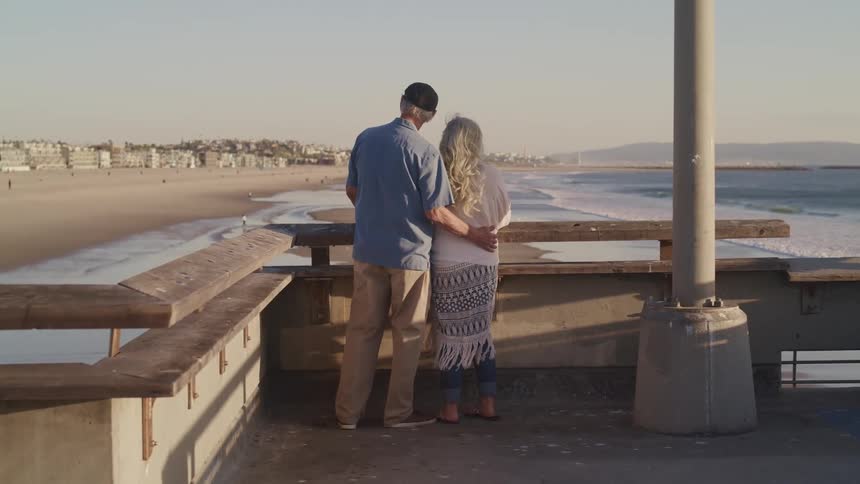 An elderly couple look out over a beach from a bridge on a sunny day