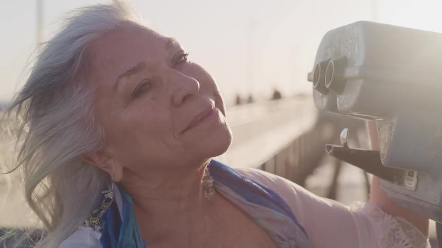 A smiling elderly woman stands next to a viewfinder on a boardwalk outdoors on a sunny day