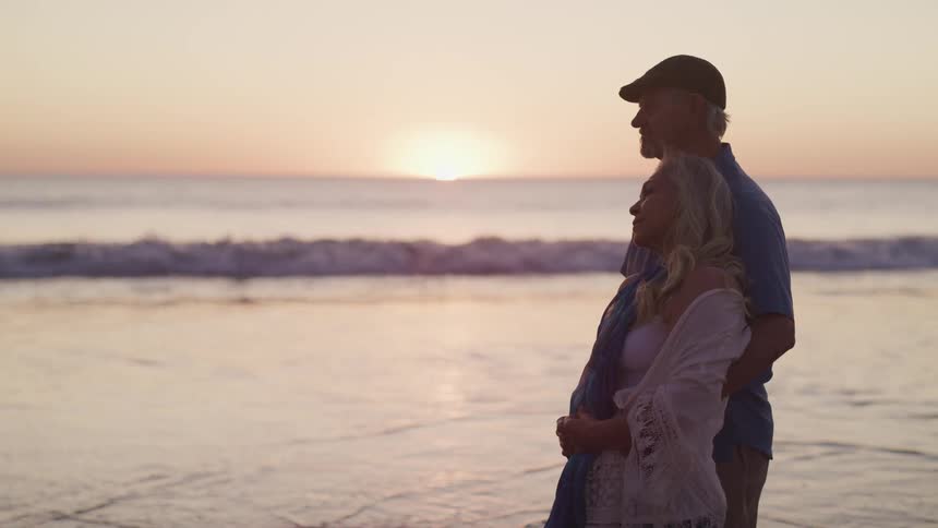 Side view of an elderly couple standing on the beach by seaside at sunset
